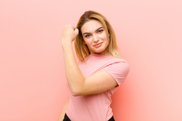 young pretty blonde woman feeling happy, satisfied and powerful, flexing fit and muscular biceps, looking strong after the gym against pink flat wall