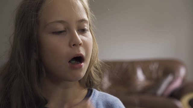 Close-up face of sick caucasian girl swallowing a green herbal pill and smiling while sitting at home. The child has fever. Concept of health, illness, sickness, common cold, treatment