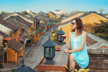Fototapeta na wymiar Tourist girl drinks coffee overlooking the old city of Hoi An, Vietnam. A young woman is enjoying the top view of the old Vietnamese city from the roof. A popular tourist destination in Vietnam