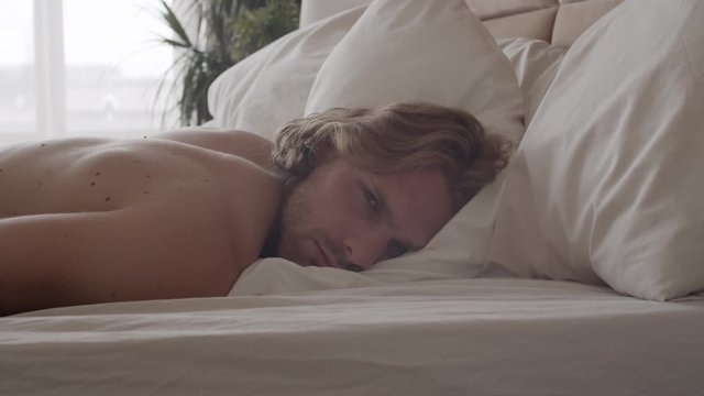 Slowmo tracking of exhausted young man lying awake in bed and looking at camera