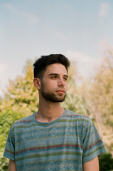 A handsome young brunette guy in a striped T-shirt stands against a background of plants and a blue...