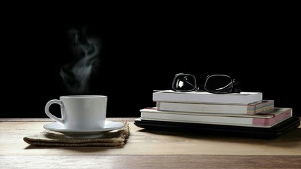 White ceramic coffee cup and eyeglasses on stacked books with laptop on wooden table in black background, coffee break concept