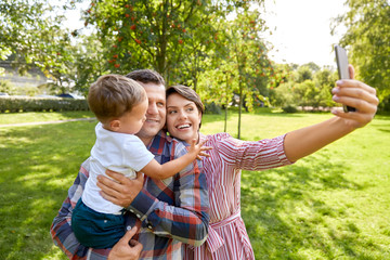 Fototapeta premium family, leisure and people concept - happy mother, father and little son taking selfie by smartphone at summer park