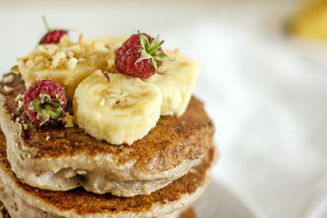 Delicious banana pancakes on white plate Stack of homemade appetizing fritters with sliced banana and fresh raspberries served on blurred white background