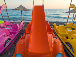Pink, orange and yellow pedal (paddle) boats - pedalo with slides, parked on a sandy beach after sunset. Straw umbrellas, calm blue sea water and clear sky in the background. In Canj (Čanj) Montenegro