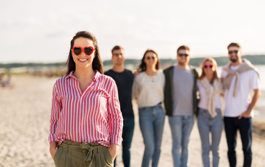 Obraz premium friendship, valentine's day and people concept - happy woman in heart-shaped sunglasses with group of friends on beach in summer