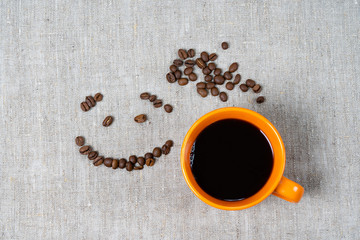 A mug of hot black coffee, coffee beans in the form of a smile and a heart on a fabric background.