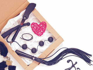 Various sex toys (leather lash, love balls, vibrator and others) are on white wrapping paper in a cardboard box. Among them is a red wicker figure of the heart. Valentine's Day Greeting Card