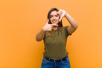 young pretty woman feeling happy, friendly and positive, smiling and making a portrait or photo frame with hands isolated against orange wall