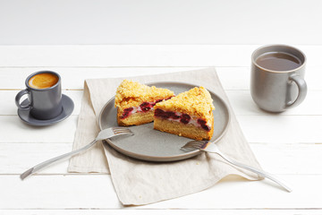 Two pieces of homemade cherry pie with crumbs on top and two cups with coffee and tea on white wooden table.
