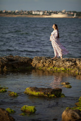 Fototapeta na wymiar Beautiful pregnant woman at sea standing on a rock in a beautiful long dress and holding her stomach. Concept: rest for pregnant women.