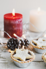 Mini mince pies on wooden table served with red and white candle, pine cones and decorative lights. Christmas and New Year mood. Festive table