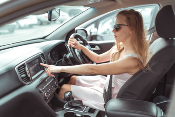 girl in car selects application on touch screen, navigation application on Internet, strapped holds wheel, left-hand traffic, wheel on right side. Woman in summer sunglasses in city.