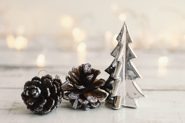 Mini christmas tree of silver color and pine cones on wooden table, Christmas lights as background. Winter decor