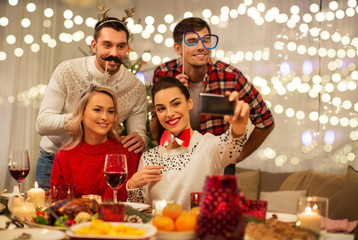 holidays and celebration concept - happy friends with party props taking selfie by smartphone at home christmas dinner