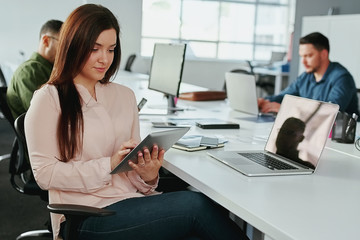 An attractive young smiling businesswoman using digital tablet with colleagues working at desk in background at office - startup office