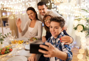 Obraz premium celebration, holidays and people concept - happy family having dinner party at home and taking selfie by smartphone