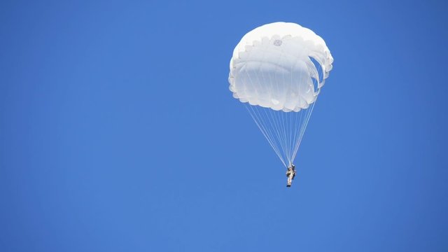 Military man in a white uniform landing on a white round parachute in a blue sky