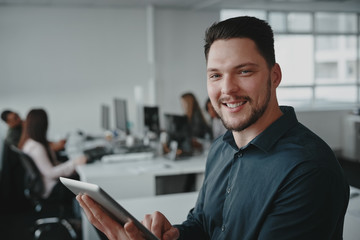 Portrait of confident smiling young businessman holding digital tablet in hand with his colleagues working at desk in background