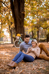 Fototapeta premium Young couple sitting on ground and using mobile phone in autumn park