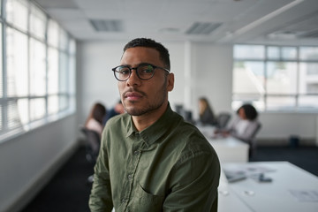 Portrait of a serious young professional businessman wearing eyeglasses looking at camera while colleague at background - obrazy, fototapety, plakaty