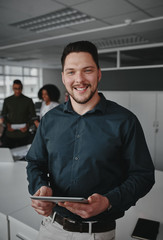 Portrait of a successful smiling young businessman holding digital tablet in hand looking at camera while coworkers having conversation on background in modern office