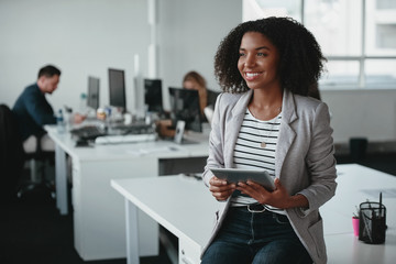 Happy thoughtful young businesswoman with digital tablet in hand smiling and looking away in front of colleague at background