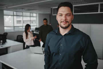 Portrait of a professional young businessman smiling and looking at camera while other business people talking in background at office