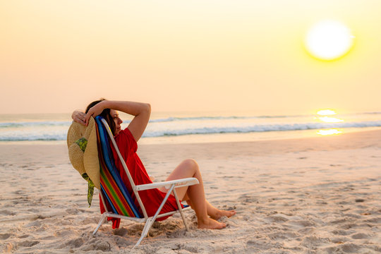 Model holding sun hat sitting on beach chair relaxing during sunset.