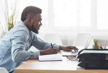 Black millennial employee using laptop in office and taking notes