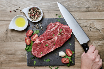 Top view of a hand with kitchen knife preparing beef steak and seasoning on wooden table