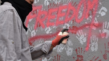 A hooded activist makes a protest graffiti on a wall