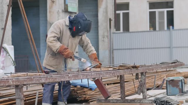 A worker at construction site is welding iron pipe with electrode in a protective uniform, orange gloves and black helmet. Sparks and smoke are allocated.