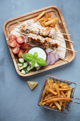 Fototapeta na wymiar Wooden serving tray with greek chicken souvlaki, tzatziki and french fries. Above view on a grey concrete background