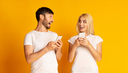 Excited Couple Talking Holding Smartphones Standing Over Yellow Background