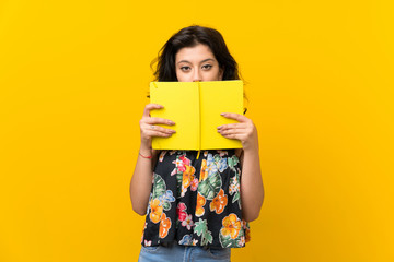 Young woman over isolated yellow background holding and reading a book