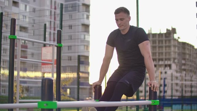Sporty man in black clothes performs lifting legs on parallel bars in slow motion against the background of buildings in the city Park