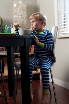 A cute little boy in striped pajamas eating cereal for breakfast at the dining room table