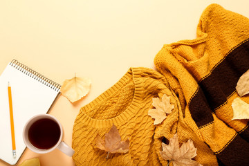 Woolen sweaters with cup of tea, notepad and autumn leafs on beige background