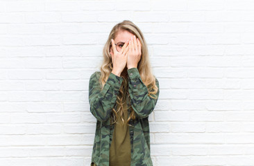 young blonde woman feeling scared or embarrassed, peeking or spying with eyes half-covered with...