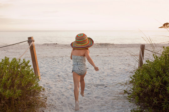A cute little girl running towards the beach in a big straw hat 