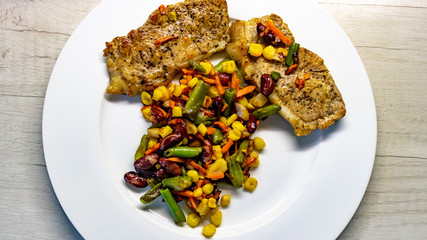 Mix of vegetable with fried meat on white plate of the light table. Mix of vegetable containing carrots, peas, beans and corn. Fried pork on a plate
