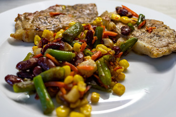 Mix of vegetable with fried meat on white plate of the light table. Mix of vegetable containing carrots, peas, beans and corn. Fried pork on a plate