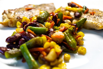 Mix of vegetable with fried meat on white plate of the light table. Mix of vegetable containing carrots, peas, beans and corn. Fried pork on a plate