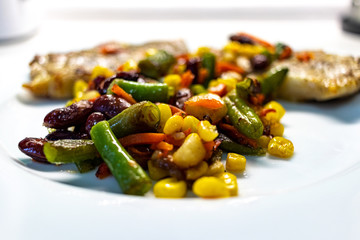 Mix of vegetable with fried meat on white plate of the light table. Mix of vegetable containing carrots, peas, beans and corn. Fried pork on a plate