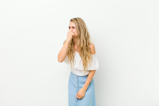 young blonde woman feeling disgusted, holding nose to avoid smelling a foul and unpleasant stench against white wall