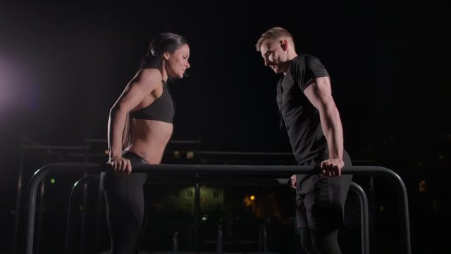 In slow motion, a man and a woman perform push-UPS on parallel bars together in the dark in a city Park. Night training