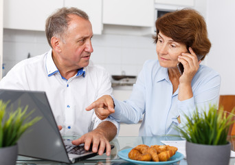 Smiling mature family couple with phone using laptop at kitchen table