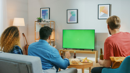 Fototapeta premium Diverse Group of Friends Sitting on a Couch at Home, Watch Green Chroma Key Screen TV while Eating Snacks and Drinking Beverage. Young People Having Fun at Home.
