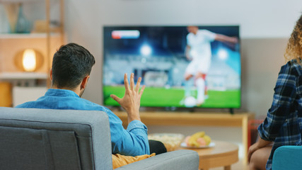 At Home Young Couple Watches Tense Moment of Soccer Match on TV, They Worry, Guy Trying to be Patient and Aggressively Showing Palm. Back View Shot.